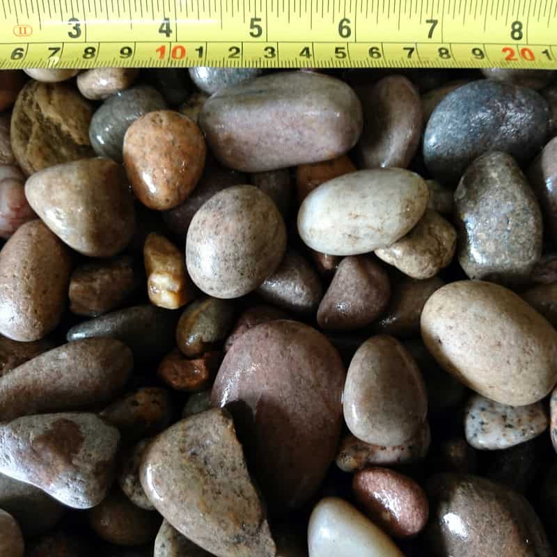 A set of smooth, oval-shaped Scottish Highland Pebbles (20-30mm) in shades of brown, gray, and beige with a yellow measuring tape above them showing measurement increments.