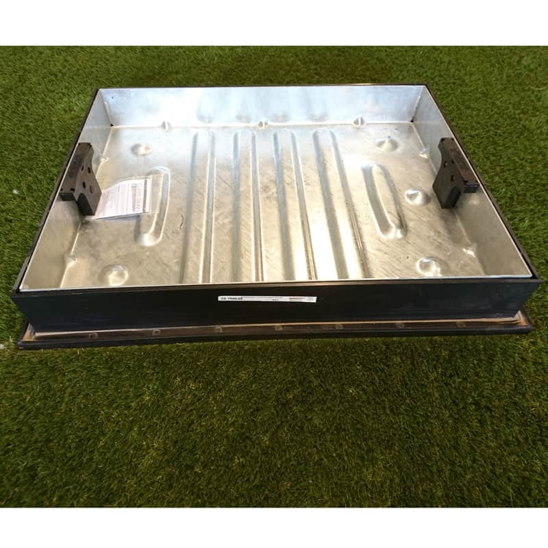 A 600x450x80mm Recess Cover & Frame with raised edges and brackets on opposite sides rests on green turf, holding a piece of paper. The surface has a ribbed texture.