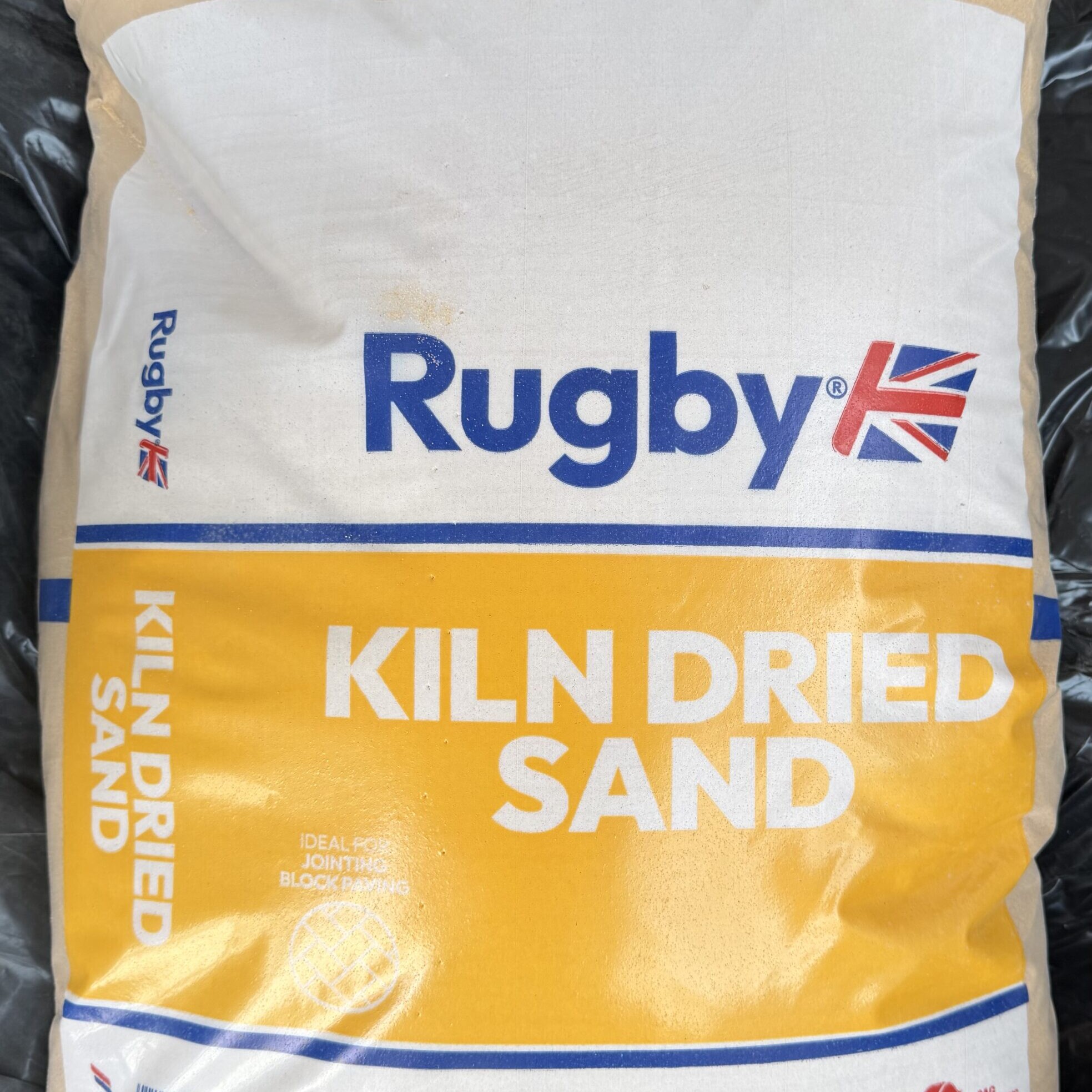 A 25kg Bag Kiln Dried Sand with yellow and white packaging, blue and red text, Union Jack flag, and CEMEX logo. The bag states it is ideal for jointing block paving.