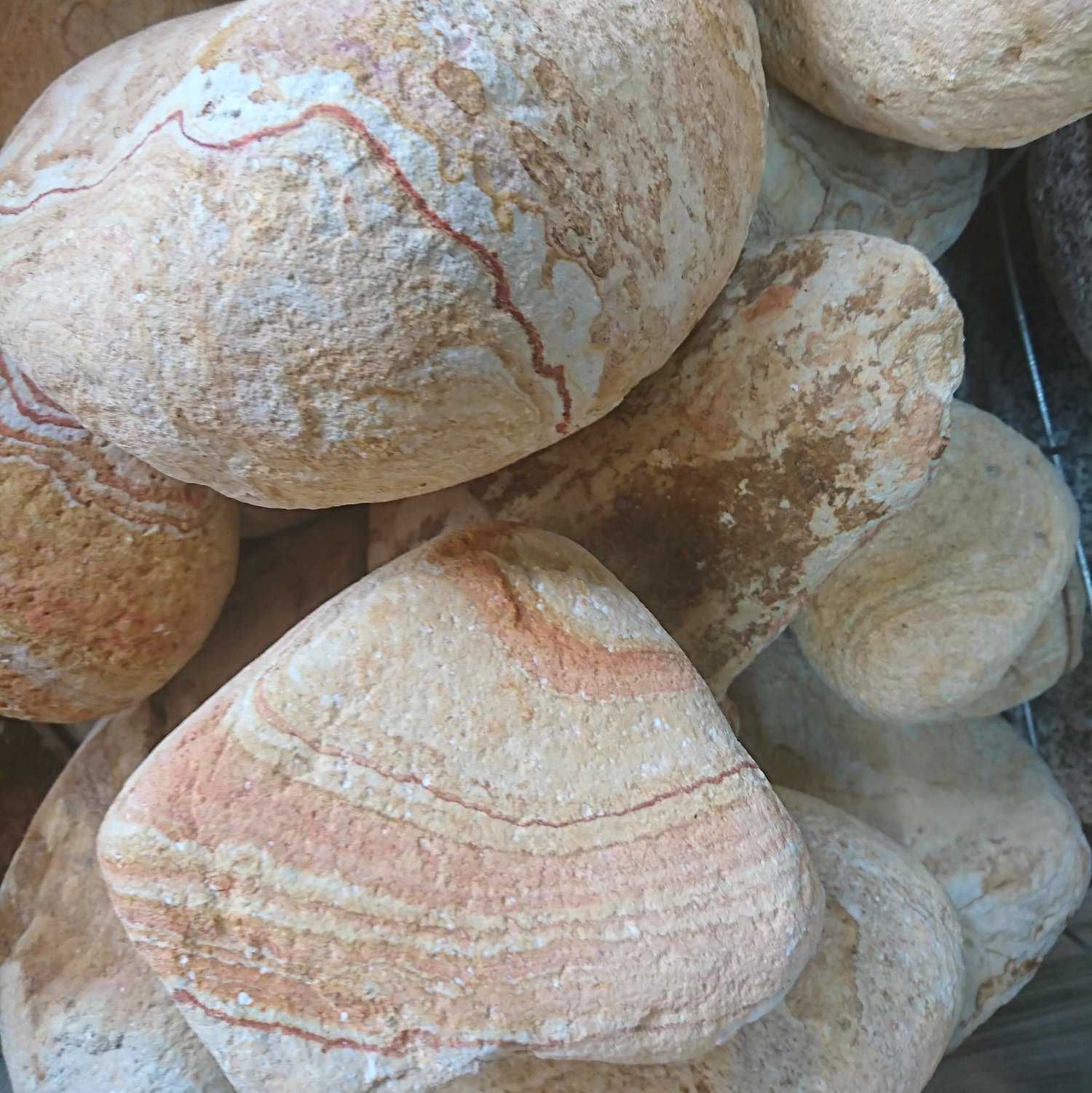 A close-up of 200-300 Riviera Marbled Boulders shows large, smooth stones with layered patterns in brown, beige, and red hues. These stones feature a naturally weathered texture as they are artistically piled.