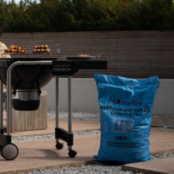A black barbecue grill with food on a patio stands beside a large blue bag labeled "Restaurant Grade BBQ Lumpwood Charcoal - 12kg," set against a modern garden backdrop for perfect grilling.