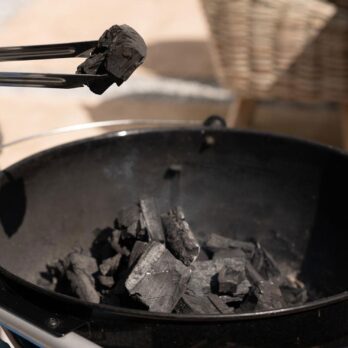 A close-up of tongs holding a piece of Homefire BBQ Lumpwood Charcoal 4Kg over a grill filled with more pieces, with a wicker basket blurred in the background.