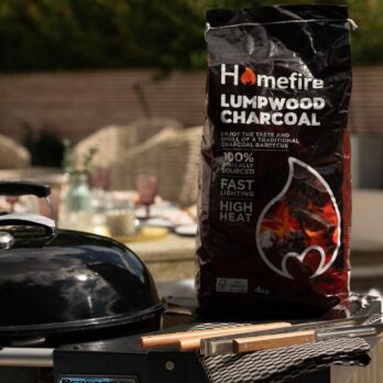 A 4kg bag of Homefire BBQ Lumpwood Charcoal is placed next to a barbecue grill outdoors, with blurred table and chairs in the background, setting the scene for a charcoal BBQ gathering.