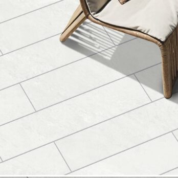 A wicker chair with a beige cushion sits on a white, rectangular-tiled floor. Sunlight highlights the clean, modern look of the Premier Boarder Porcelain Tile White 900 x 150 x 20mm beneath it.