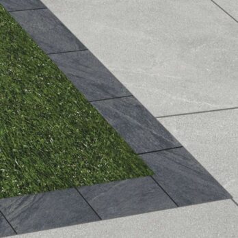 A triangular patch of green grass is edged with Premier Boarder Porcelain Tile Anthracite 900 x 150 x 20mm and surrounded by large, light gray concrete pavers.