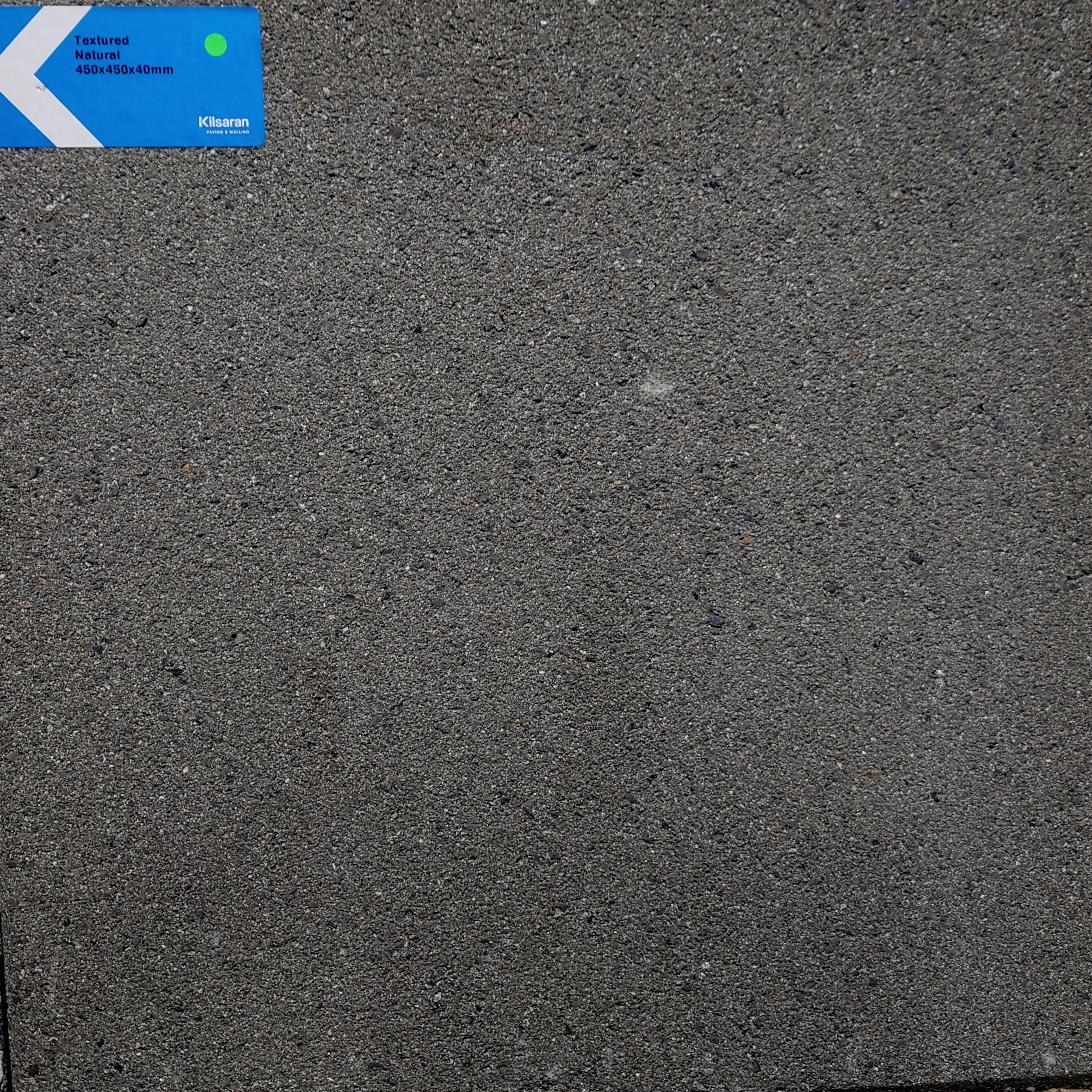 A close-up of a textured, gray Kilsaran Textured 450 x 450 x 40mm Natural Flag Stone slab with a small blue and white label in the top left corner displaying product details for patios and footpaths.