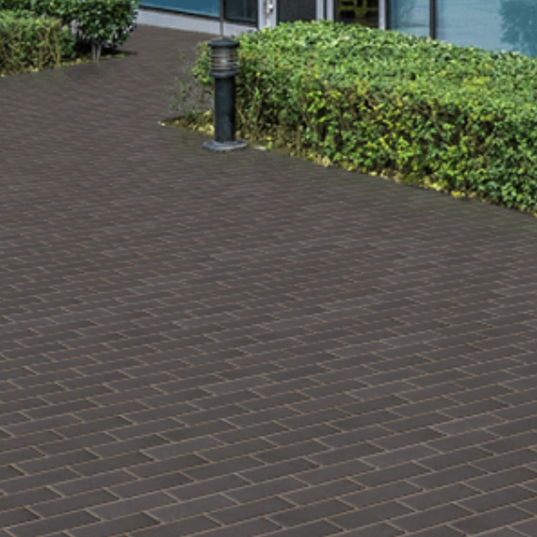 A courtyard paved with Kilsaran Slane 50mm Driveway Charcoal, bordered by a green hedge and a modern building with blue window panes. A black outdoor lamp is positioned near the hedge.