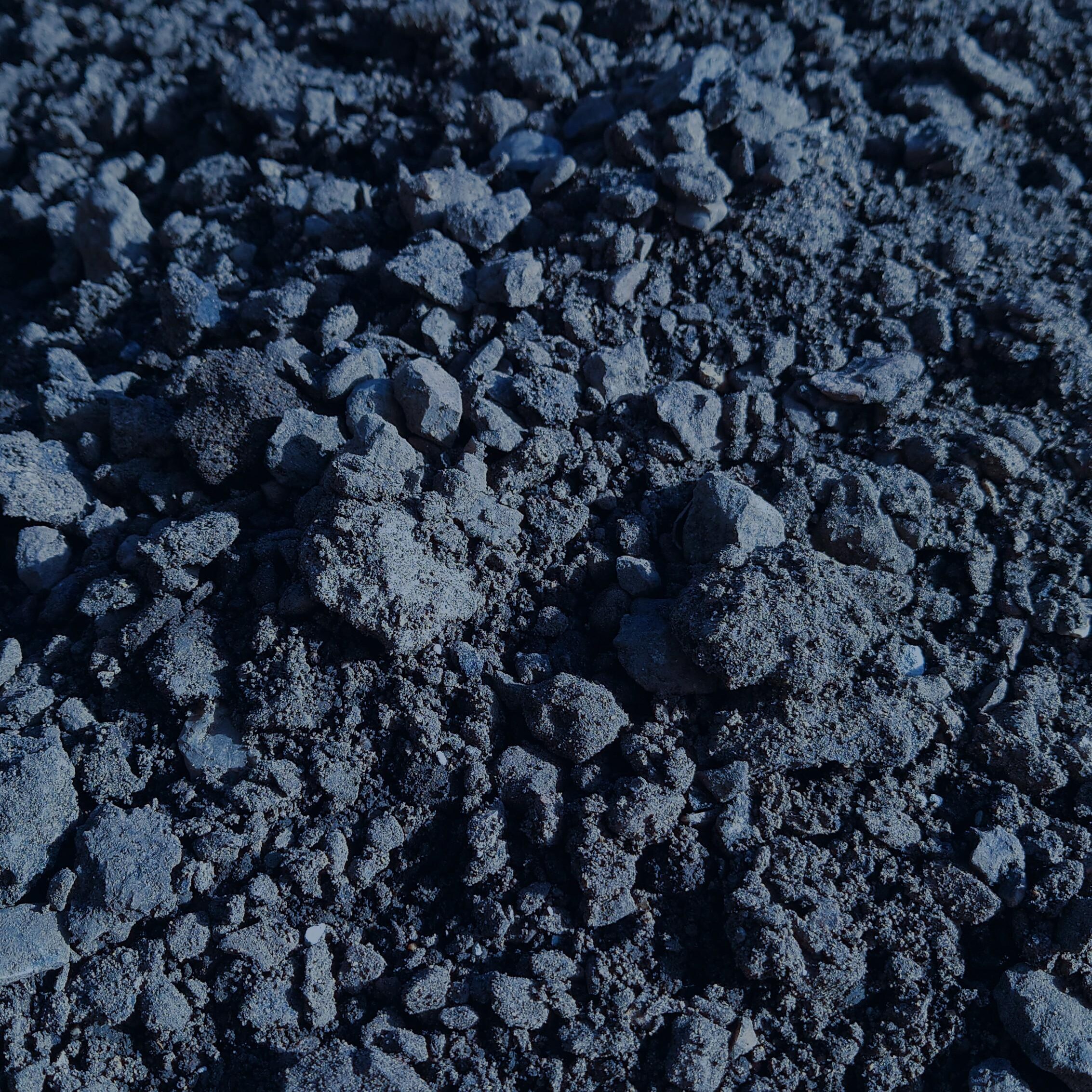 A close-up of Road Planings Graded - Loose or Bagged, showing a rough, uneven surface with dark grey soil, small stones, and scattered road planings scaplings that highlight the varied textures and clumps throughout.