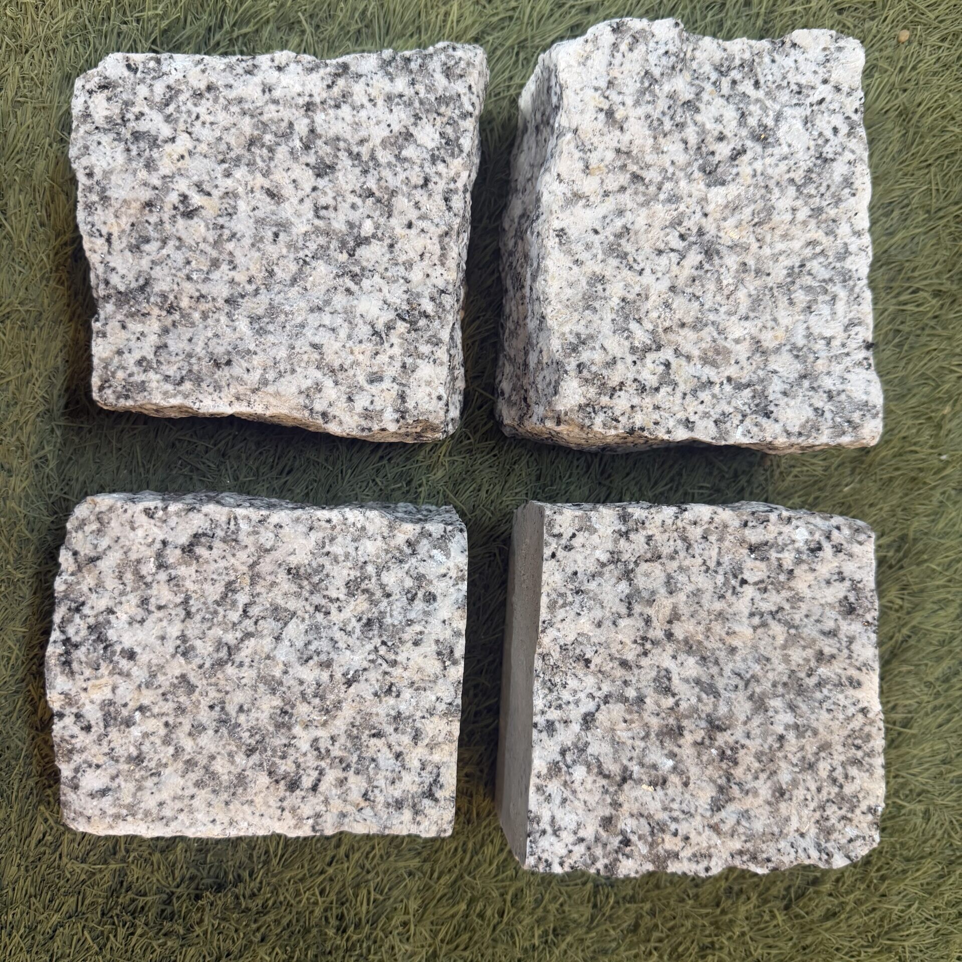 Four Granite Setts Hand Cut 100x100x40/60mm, each square with rough edges, are arranged in a two-by-two grid on a textured green surface.
